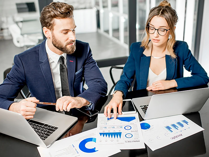 A man and a woman in business attire sit at laptops and look at statistics