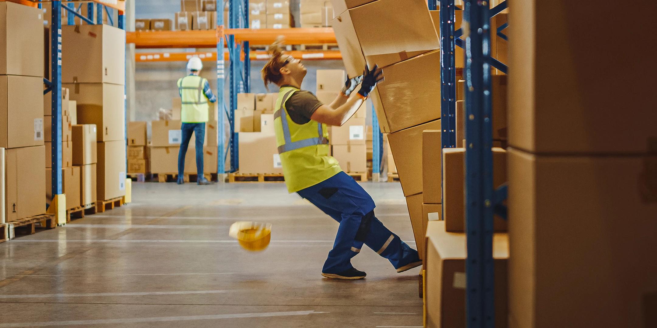 Two men working in a warehouse. The man in the foreground is trying to pick up a stack of boxes that has just fallen over. His hard hat falls to the ground.